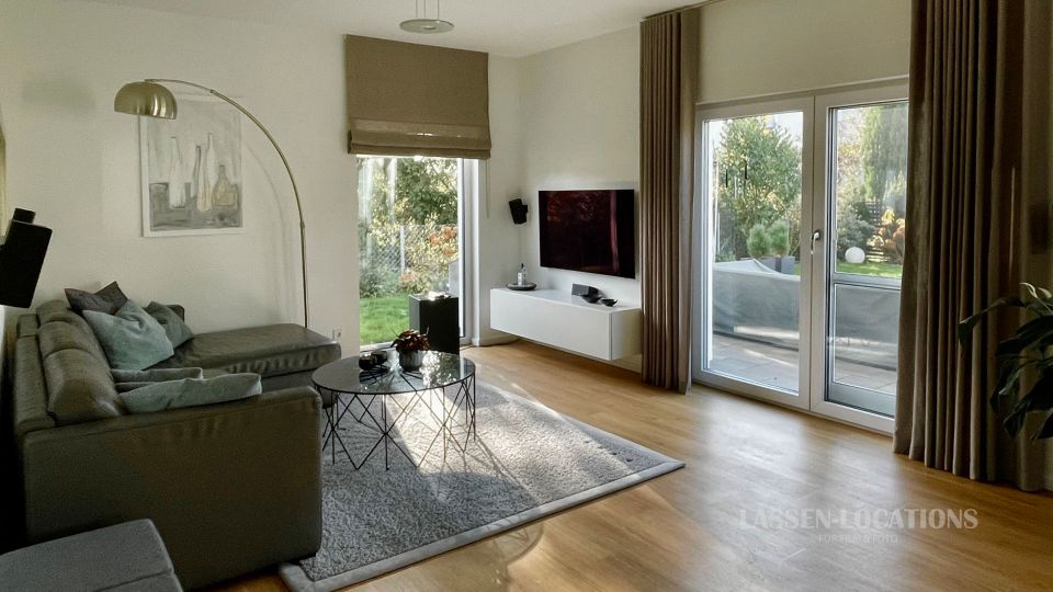 Modernes Wohnzimmer mit grauem Ecksofa, rundem Couchtisch auf Teppich und wandmontiertem Fernseher über einem schwebenden Sideboard. Große Glas-Terrassentüren und ein schmales Fenster mit Vorhängen lassen Tageslicht herein und geben den Blick in einen grünen Garten frei.