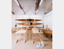Werkstatt mit Holzkajaks: Mehrere handgefertigte Holzkajaks hängen an einer weißen Wand auf Regalen. Im Vordergrund stehen Werkbänke mit Werkzeugen und Baumaterialien, der Boden ist mit Sperrholzplatten ausgelegt, und die Decke zeigt Leuchtstofflampen und freiliegende Strukturen.