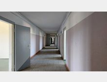 Langer, schlichter Flur eines Gebäudes mit hellvioletten Wänden und teilweise offenen Türen auf beiden Seiten, grauem Boden aus Fliesen und einer klaren Sichtachse bis zum Ende des Gangs.