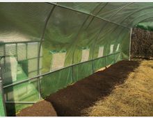 Ein Gewächshaus mit grünem Netzstoff über einer Metallrahmenkonstruktion, mit kleinen rechteckigen Lüftungsöffnungen. Im Inneren sind frisch vorbereitete Beete mit Erde und eine Fläche mit Stroh zu sehen.