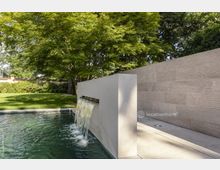 Moderner Garten mit rechteckigem Wasserbecken und einer rechteckigen Wand aus hellem Stein, aus der ein kleiner Wasserfall fließt. Im Hintergrund sind eine Rasenfläche, hohe Bäume und eine Mauer mit Büschen zu sehen.