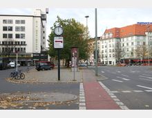 Städtische Straßenkreuzung mit breiten Fahrbahnen, einem roten Radweg und herbstlich gefärbtem Laub auf dem Boden. Links ein mehrstöckiges Gebäude mit der Aufschrift „Die Wühlmäuse“ und rechts eine Reihe von Wohn- und Geschäftshäusern mit roten Dächern.