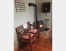 Gemütliche Ecke eines Raumes mit einem brennenden Holzofen, einem schwarzen Ofenrohr, einer Backsteinunterlage und einer hölzernen Sitzbank mit rot-weiß karierten Kissen. An der Wand hängen ein dekoratives Schild mit Schriftzug und ein Regal mit kleinen Fächern.