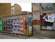 Holzwand und Gebäudefassade in einer urbanen Umgebung, bedeckt mit bunten Plakaten für Konzerte und Veranstaltungen. Umgebende Wände zeigen Graffiti, mit einem Hintergrund aus Altbauten und einem Gehweg im Vordergrund.