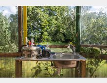 Eine einfache Spüle in einem Außenbereich mit Blick auf grüne Bäume und Wiese. Der Bereich ist von transparenten und hölzernen Paneelen umgeben, und auf der Spüle liegen Geschirr, Putzutensilien und eine kleine Bürste.