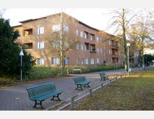 Mehrfamilienhaus in einem hellbraunen Farbton mit mehreren Balkonen, umgeben von Bäumen und Sträuchern. Im Vordergrund befindet sich ein gepflasterter Weg mit grünen Parkbänken sowie Grasflächen und spärlich belaubten Bäumen.