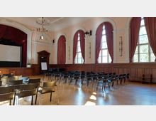 Ein historischer Saal mit hohen Decken, großen Bogenfenstern und roten Vorhängen. Vorne befindet sich eine Bühne mit rotem Samtvorhang und einer Leinwand, während der Raum mit schwarzen Stühlen in Reihen bestuhlt ist.