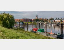 Flusslandschaft mit einem kleinen Hafen, Booten und einer Anlegestelle; im Vordergrund eine grasbewachsene Böschung und ein Schiff mit roten und grünen Bojen, im Hintergrund eine Altstadt mit roten Ziegeldächern und einer Kirchturmspitze.