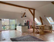 Offener Dachgeschossraum mit schrägen Decken, Holzträgern und Terrakottafliesen. Der Raum enthält eine moderne, weiße Kücheninsel, einen kleinen Treppenbereich mit Holzgeländer, große Dachfenster, eine Glastür zu einem Balkon und minimalistische Einrichtung.