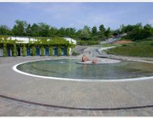 Runder Brunnen mit flachem Wasser und einer abstrakten Skulptur in der Mitte, umgeben von gepflasterten Wegen und begrünten Wänden mit rankenden Pflanzen; im Hintergrund eine hügelige Landschaft mit Treppen und Bäumen.