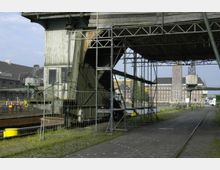 Industrielles Hafengelände mit einer alten Metallkonstruktion, teils von Gerüsten umgeben, im Vordergrund. Im Hintergrund sind historische Backsteingebäude, ein Wasserkanal und moderne Bauten sichtbar.