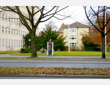 Eine städtische Szene mit einem kleinen Park, in dem ein moderner Telefonhäuschen steht. Im Hintergrund sind ein mehrstöckiges Verwaltungsgebäude und ein beiges Wohnhaus mit großen Fenstern zu sehen, umgeben von Bäumen und Sträuchern.