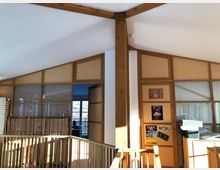 Innenaufnahme eines modernen Gebäudes mit schrägen Decken und Holzelementen. Holzgitter, halbhohe Trennwände und Glasfenster trennen Bereiche; an den Wänden hängen Poster und Bilder.