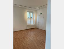 Ein leerer Raum mit hellem Laminatboden, einer weißen Wand, einem Fenster mit Vorhängen und einer weißen Tür mit Fluchtschild. Oben sind kleine Deckenspots angebracht, und unter dem Fenster befindet sich ein Heizkörper.