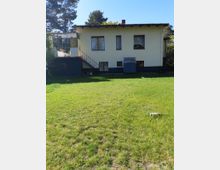 Ein zweistöckiges Wohnhaus mit gelber Fassade und Flachdach, umgeben von einem großen, grünen Rasen. Links befindet sich ein Treppenaufgang zu einem Balkon mit Überdachung, und im Hintergrund sind Bäume zu sehen.