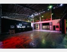 Innenansicht eines leeren Veranstaltungsraums mit hohen Decken, sichtbaren Stahlträgern und einer spiegelnden Discokugel in der Mitte. Der Raum hat eine dunkle Bar auf der linken Seite und einen offenen Durchgang auf der rechten, beleuchtet durch farbige Lichter.