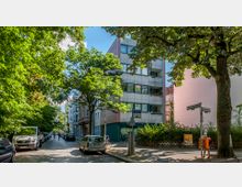 Straßenszene in einer Wohngegend mit Kopfsteinpflaster, flankiert von Bäumen und geparkten Autos. Im Hintergrund steht ein modernes Wohngebäude mit Balkonen, und Straßenschilder weisen auf eine Kreuzung hin.