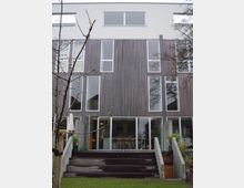 Mehrstöckiges modernes Wohngebäude mit Holzelementen an der Fassade und großen Glasfenstern. Im Erdgeschoss befindet sich eine Terrasse mit breiten Holzstufen und umgebender Begrünung.