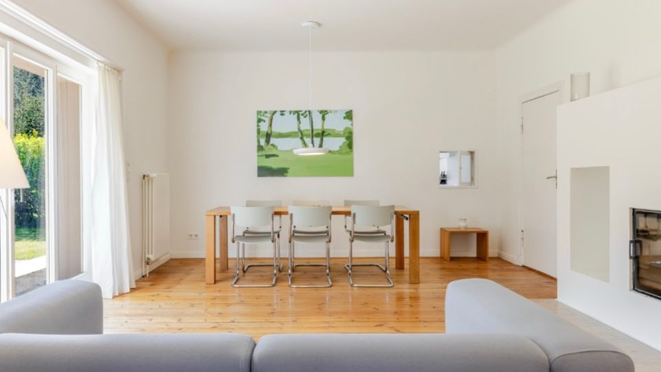 Helles Wohn- und Esszimmer mit Holzboden, großer Terrassentür mit Vorhängen links und einem Esstisch mit vier Stühlen unter einer Pendelleuchte. Rechts befindet sich ein weißer Kamin mit Nische, an der Rückwand hängt ein Landschaftsbild.