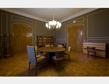 Ein klassisch eingerichteter Raum mit Holzboden, runden Holztisch und mehreren gepolsterten Stühlen in der Mitte. Der Raum besitzt zwei große Holztüren, eine Bücherwand, ein Klavier mit Stuhl sowie eine Deckenlampe mit mehreren Leuchten.