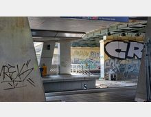 Bahnhofsumgebung mit Betonstützen unter einer Bahnüberführung, bedeckt mit Graffiti. Im Vordergrund befinden sich ein Bahnsteig mit Sitzmöglichkeiten und eine Treppe, die zu den Gleisen führt.