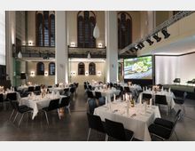 Ein großer Veranstaltungsraum in einem modernen, umgebauten historischen Gebäude mit hohen Decken und großen Bogenfenstern. Der Raum ist mit weiß gedeckten Tischen, Stühlen und Kerzen dekoriert; im Hintergrund befindet sich eine Bühne mit einem projizierten Bild und Beleuchtung.