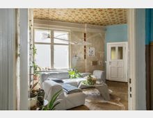 Wohnzimmer mit hellgrauen Sofas und einem modernen Couchtisch auf einem Kuhfell-Teppich, dekoriert mit Zimmerpflanzen. Die Wände sind teilweise verputzt, mit einer alten Tapete an der Decke, großen Fenstern und einer weißen Holztür im Hintergrund.