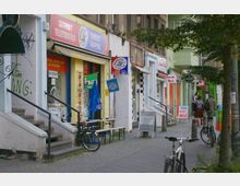 Straßenszene mit kleinen Geschäften in einer städtischen Umgebung, darunter ein Internetladen, ein Nagelstudio und weitere Gewerbe, vor denen Fahrräder abgestellt sind. Die Gebäude haben Körpereingänge, teils mit Treppen, und es stehen Bäume entlang des Gehwegs.