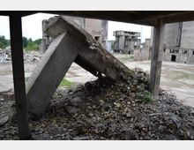 Ein verlassenes Industriegebiet mit zerfallenen Betonstrukturen und Schutthaufen. Im Vordergrund steht eine eingestürzte Betonstütze, umgeben von Trümmern und überwuchertem Gelände, während im Hintergrund verfallene Gebäude sichtbar sind.