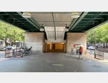Unterführung unter einer Brücke mit grüner Stahlkonstruktion und großem Betonträger. Auf der rechten Seite ist eine graffitibemalte Wand mit Fahrrädern davor, links sind weitere abgestellte Fahrräder sowie ein Elektroroller; dahinter Autos und Bäume an der Straße.