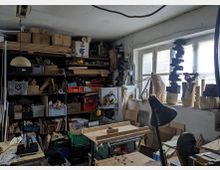 Werkstatt mit Holzwerken und Werkzeugen. Die Wände sind mit Regalen gefüllt, die verschiedene Holzmaterialien, Kartons und Werkzeuge enthalten; auf einer Werkbank stehen Maschinen und eine Tischlampe, während am Fenster Holzskulpturen arrangiert sind.