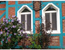 Fachwerkhausfassade aus roten Ziegeln mit türkisfarbenen Holzbalken, zwei weißen Fenstern mit Spitzbögen und einem Wappen oberhalb der Fenster; im Vordergrund ein blühender Strauch mit violetten Blüten.
