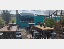 Eine Outdoor-Café- oder Barfläche mit Holztischen, Metallhockern und zahlreichen Blumenarrangements. Im Hintergrund steht ein türkisfarbener Container mit einer Theke, umgeben von Bäumen, Pflanzen und Beleuchtung.