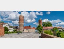 Historische Stadtansicht mit zwei runden, mittelalterlichen Backsteintürmen, die Teil einer alten Stadtmauer sind; im Vordergrund eine gepflasterte Straße mit umliegenden Häusern und vereinzelten Bäumen unter einem wolkigen, blauen Himmel.