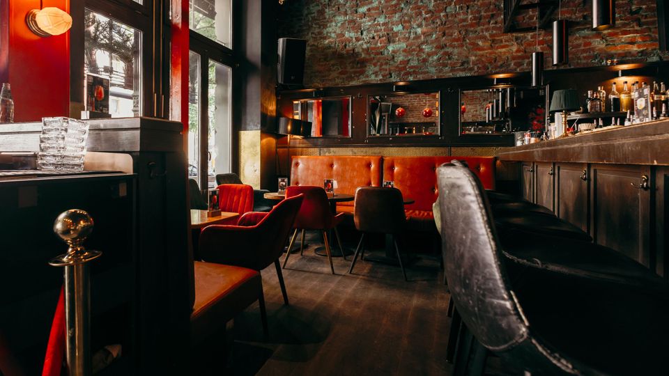 Innenraum einer stilvollen Bar mit freiliegender Backsteinwand, dunkler Holztheke mit Flaschenregal und warmen Leuchten. Vor großen Fenstern stehen kleine Tische mit roten Polstersesseln und einer gepolsterten Sitzbank entlang der Wand.