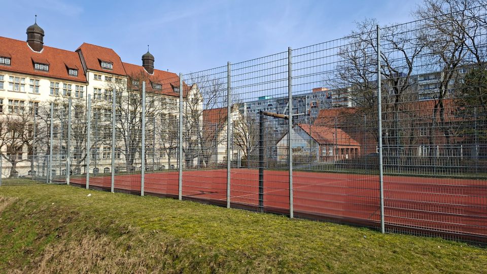 Eingezäunter Sportplatz mit rotem Kunststoffbelag und Basketballkorb, umgeben von Grasfläche und kahlen Bäumen. Im Hintergrund stehen mehrstöckige Gebäude mit roten Ziegeldächern sowie ein Hochhaus.