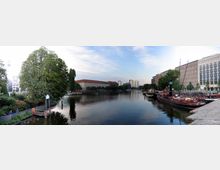 Panorama einer Flusslandschaft mit ruhigem Wasser in einer urbanen Umgebung. Links ein grüner Baum und Spazierweg, rechts Anlegestellen mit Schiffen, im Hintergrund Gebäude mit roten und hellen Dächern sowie moderne Hochhäuser.