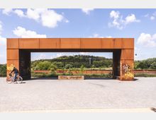 Rostfarbener Torbogen aus Metall in einer urbanen Umgebung, mit Graffiti verziert, durch den man auf eine bewaldete Landschaft mit Hügeln und einer Seilbahn im Hintergrund blickt; im Vordergrund ein gepflasterter Platz mit einem Radfahrer.