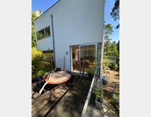 Moderne Hausfassade mit großer Schiebetür aus Glas, die auf eine Holzterrasse führt. Auf der Terrasse stehen ein runder Metalltisch, Stühle und Pflanzkübel; seitlich verläuft ein Glasgeländer mit Blick ins grüne, baumbestandene Umfeld.