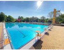 Ein Freibad mit einem großen, rechteckigen Schwimmbecken, markiert durch Schwimmbahnen und Startblöcke am Rand. Im Hintergrund sind ein gelber Sprungturm, eine rote Wasserrutsche und grüne Bäume zu sehen; der Himmel ist klar und sonnig.