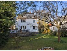 Rückansicht eines weißen, mehrstöckigen Wohnhauses mit schrägem Ziegeldach und Dachgauben, davor eine breite Treppe zur Terrasse. Im Vordergrund liegt ein großer Rasen mit Bäumen, Sträuchern und einem kleinen Holzstapel, nebenan ist ein weiteres Wohnhaus zu sehen.