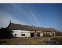 Langes, niedriges Wirtschafts- oder Scheunengebäude mit großem Satteldach und gemischter Fassade aus weiß verputzten und beige-grauen Bereichen, teils mit großen Toröffnungen und Fenstern. Davor liegt eine verwilderte Wiese mit einigen Holz- und Baumaterialresten, im Hintergrund stehen kahle Bäume unter blauem Himmel mit dünnen Wolkenschleiern.