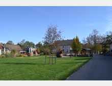 Grüne Parkanlage mit einem großen Rasenbereich, einem kleinen Baum in der Mitte und einer dekorativen Skulptur in Form eines schrägen Gefäßes. Im Hintergrund befinden sich Gebäude mit Ziegeldächern, überdachte Sitzbereiche und ein gepflasterter Weg sowie Bäume mit herbstlichem Laub.