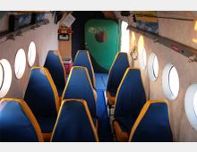 Innenraum eines kleinen Flugzeugs mit zwei Sitzreihen, die aus blauen Sitzen mit gelben Rändern bestehen. Die Wände sind beige mit runden Fenstern und einer grünen Tür im hinteren Bereich.