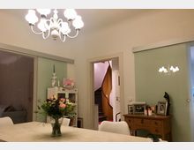 Ein Esszimmer mit einem hellen Kronleuchter, einem Holztisch mit Blumenstrauß in der Mitte und weißen Stühlen. Im Hintergrund sind eine Tür, die zu einer Treppe führt, ein Sideboard mit gerahmten Fotos und eine Glas-Schiebetür zu sehen.