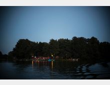Eine Abendaufnahme von einem See mit ruhiger Wasseroberfläche, umgeben von Bäumen. Am Ufer ist ein beleuchtetes Gebäude oder Restaurant mit Lichterketten und reflektierenden Lichtern im Wasser zu sehen.