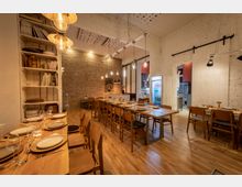 Innenraum eines modernen Restaurants mit rustikalem Stil. Der Raum hat eine Ziegelwand, Holztische und -stühle, helle Hängeleuchten sowie eine offene Küche; ein Regal mit Geschirr und ein Kühlschrank mit Getränken sind ebenfalls sichtbar.