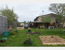Ein ländlicher Garten mit mehreren Baumbeeten, offenen Rasenflächen und Hochbeeten, umgeben von vereinzelt stehenden Bäumen. Im Hintergrund steht ein einfaches Gebäude mit einer Veranda und einer gehissten Flagge auf dem Dach.