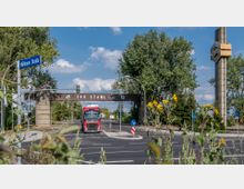 Eine Straßenansicht mit dem Eingangstor der Firma "EKO Stahl GmbH", darauf ein Schriftzug und ein Logo. Im Vordergrund fährt ein roter Lkw, umrahmt von Grünpflanzen und gelben Blumen, im Hintergrund sind Bäume und ein hoher Industrieschornstein zu sehen.