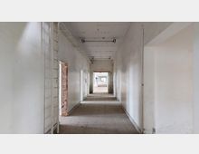 Ein leerer, schmaler Flur in einem verfallenen Gebäude mit weißen, teils beschädigten Wänden, sichtbarem Mauerwerk an einer Türöffnung und einer hell beleuchteten Fläche im Hintergrund.